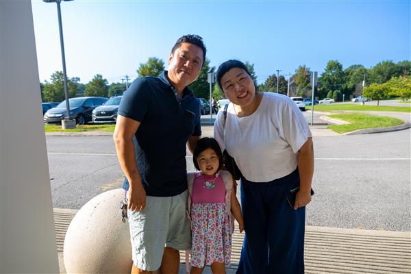mom, dad and girl student smiling in front of the parking lot
